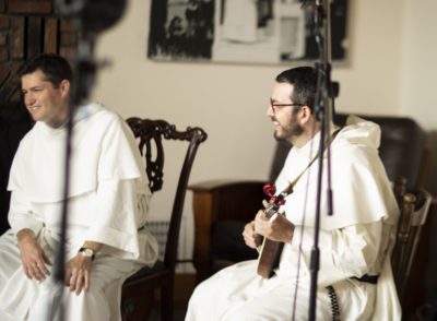 Two Dominicans sit in chairs, smiling, one holding a mandolin, the view partially obscured by microphone stands